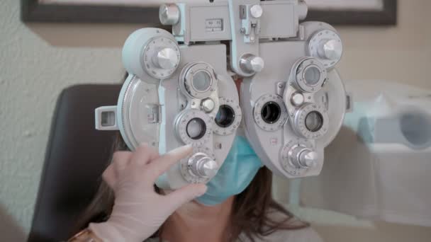 Pomiar wzroku u młodej kobiety z optycznym phoropterem. Kobieta robi test wzroku. Dziewczyna i lekarz w maskach w pandemii. Zamknij się. — Wideo stockowe