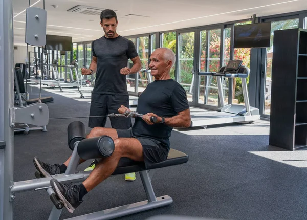 Starszy mężczyzna, atletyczna budowa ciała ćwiczy na urządzeniu treningowym, z trenerem fitness. Na siłowni. — Zdjęcie stockowe