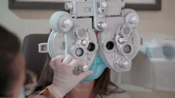 Pomiar wzroku u młodej kobiety z optycznym phoropterem. Kobieta robi test wzroku. Dziewczyna i lekarz w maskach w pandemii. Zamknij się. — Wideo stockowe