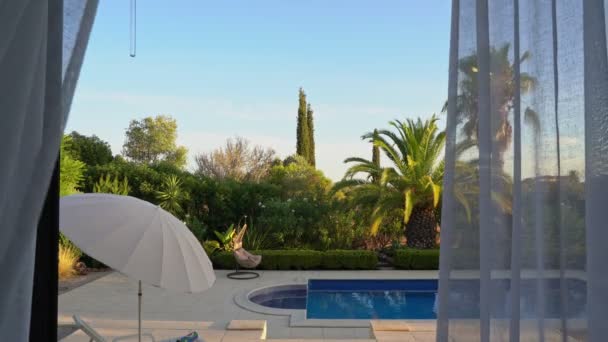 Vista desde la moderna sala de estar con cortinas soplando en el viento revelando un balcón y una piscina exterior. Y jardín tropical. — Vídeo de stock