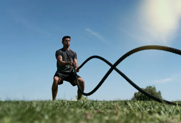 Atleta masculino ao ar livre no parque, exercícios de resistência, usando uma corda grossa, para cross fitness. Europeu no verão — Fotografia de Stock