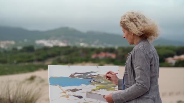 Femme européenne d'âge moyen, artiste, peint un paysage marin avec une vue originale sur la mer. Mouvement lent — Video