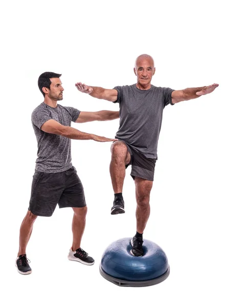 Un anciano haciendo un ejercicio de equilibrio, en una pierna, en una pelota hemisférica. Con la ayuda de un entrenador de fitness. Sobre un fondo blanco aislado. — Foto de Stock
