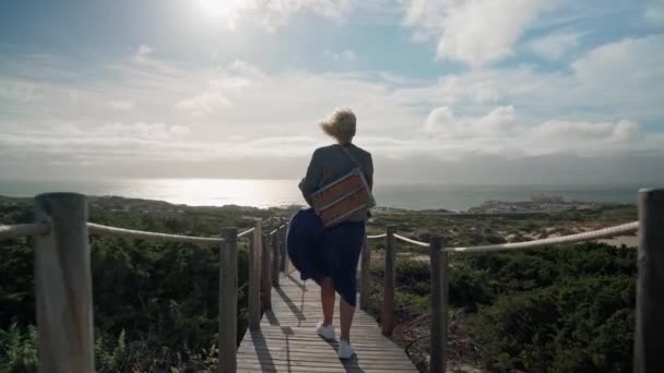Una artista europea de mediana edad con un caballete en el hombro camina hacia el océano. Puente de madera. — Vídeos de Stock