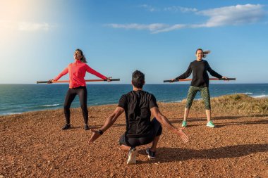 Orta yaşlı erkek ve kadın çalıştırıcı okyanus kıyısında fitness dersleri veriyor. Esnemek için jimnastik sopaları kullanıyor. Güneşli bir gün.