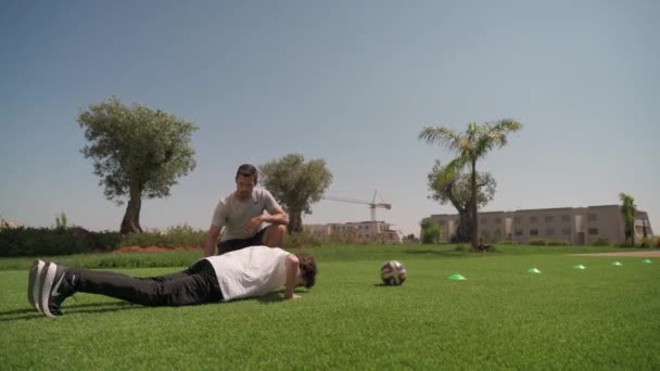 Ein Mann mittleren Alters trainiert mit einem jungen Mann im Park. Liegestütze. Laufen mit dem Ball im Zickzackkurs. — Stockvideo