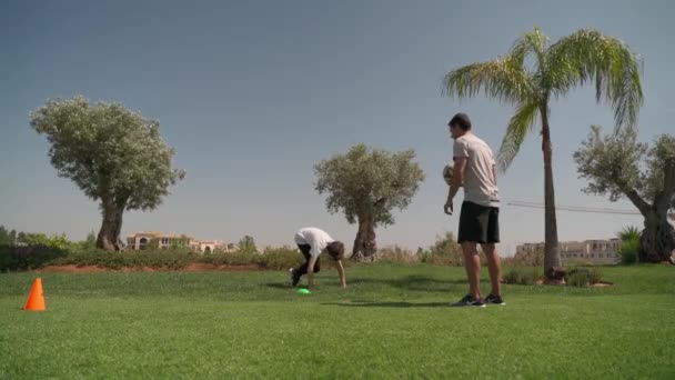 Ein junger Mann auf der grünen Wiese, der mit einem Trainer trainiert, um Fußball zu spielen. Laufen mit Hindernissen für Koordination und Schnelligkeit. — Stockvideo