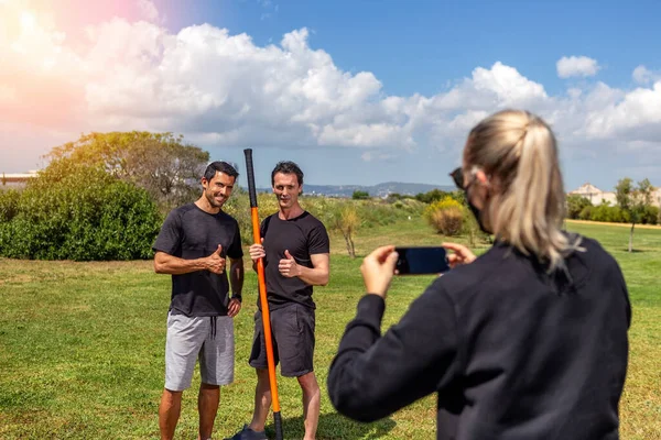 Fitness trainer e un cliente posano per una giornalista donna, sparando su uno smartphone. Giornata di sole. Con un bastone da ginnastica. estate in giardino. — Foto Stock