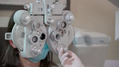 Optik foropteri olan genç bir kadının görme ölçüsü. Göz testi yapan bir kadın. Kız ve maskeli doktor bir salgın içindeler. Kapat.