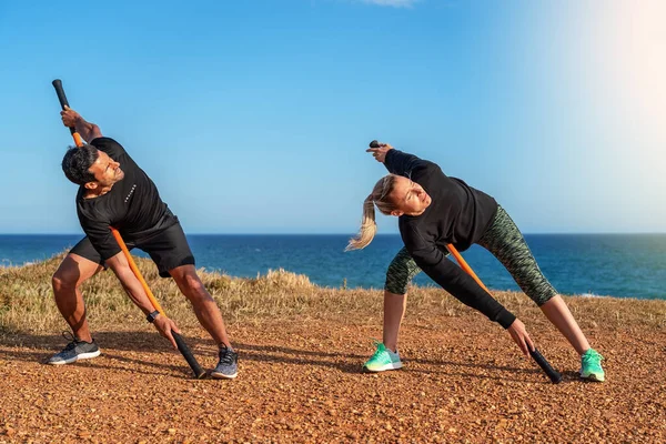 Ein Mann und eine Frau mittleren Alters auf dem Meer machen ein Warm-up. Fitnesskurse. Mit Gymnastikstöcken zum Dehnen. Sonniger Tag. — Stockfoto