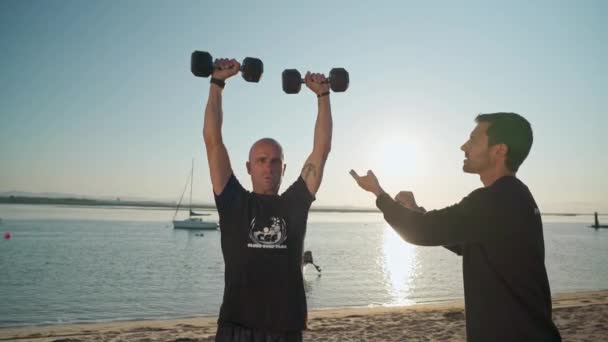 Un athlète sur la plage, s'entraîne avec des haltères et reçoit des instructions d'un entraîneur de fitness personnel. Pour une compétition de surf. Portugal Faro 2021 15 mars — Video