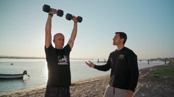 Een sporter op het strand, traint met halters en krijgt instructies van een personal fitness trainer. Voor een surfwedstrijd. Portugal Faro 2021 maart 15 — Stockvideo