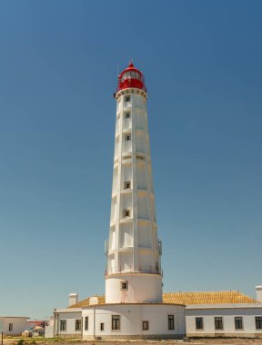 Mavi gökyüzüne karşı kırmızı kapaklı beyaz bir deniz feneri. Kumlu bir plajda. Boşluğu kopyala