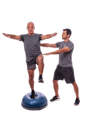 Yaşlı bir adam denge egzersizi yapıyor, tek bacakla, yarım küre topuyla. Bir fitness eğitmeninin yardımıyla. İzole edilmiş beyaz bir arkaplanda.