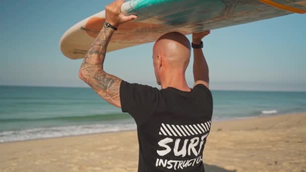 Manlig surfartränare instruktör, med tatueringar, bär en bräda på huvudet mot havet. Portugal Faro 2021 mars 15 — Stockvideo
