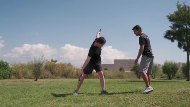 Um treinador de fitness com um cliente, em um parque em um prado verde, faz alongamentos com a ajuda de um pólo de ginástica, pau. Portugal Faro 2021 15 de Março — Vídeo de Stock