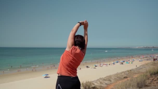 Вид сзади, женщины среднего возраста, ожирение, во время занятий гимнастикой и разогрева. На берегу моря с видом на пляж. Медленное движение — стоковое видео