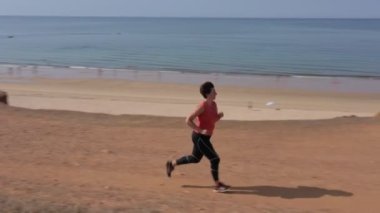 Orta yaşlı bir kadın, kilolu, kardiyo koşusu yapıyor, kumsalda, kilo vermek için. Arka planda deniz manzarası.