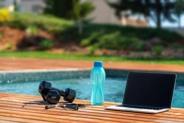 Fitness concept image of a dumbbell, a jump rope, a bottle of water and a computer for online workouts near the pool. Close up, outside. — Stock Photo, Image