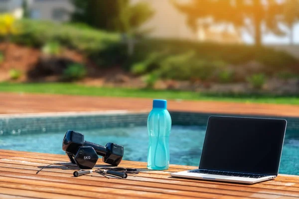 Imagem de conceito de fitness de um haltere, uma corda de salto, uma garrafa de água e um computador para exercícios online perto da piscina. Fechar, lá fora. — Fotografia de Stock