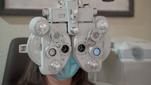 光フォロプターを持つ若い女性への視力測定。目の検査をしてる女性。パンデミックのマスクの少女と医者だ。閉じろ! — ストック動画