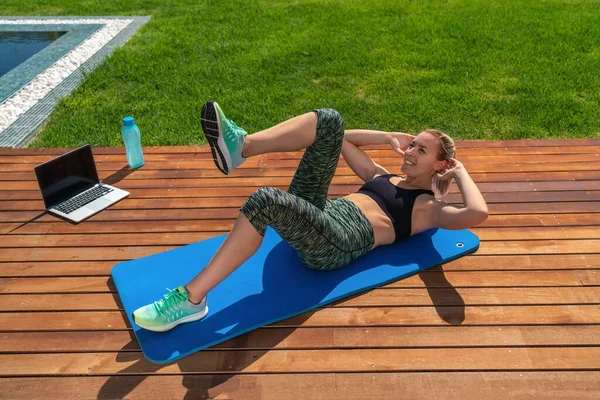 Krásná evropská dívka, provádí cvičení pro břicho, odlehlé lekce, fitness trenér na notebooku. V parku s bazénem.na trávě. — Stock fotografie