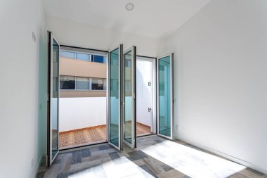 Açık güneş ışığı alan balkon ve minimalist tasarım elementleri, parlak cam kapılar, ahşap döşemeli ve temiz beyaz kaplamalı balkona açılan oda.