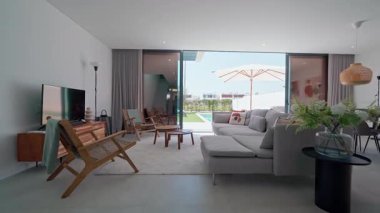 Doğal ışık modern bir oturma odasını aydınlatıyor, rahat mobilyaların altını çiziyor, büyük bir kanepe ve cam kapılardan yüzme havuzu manzarası.