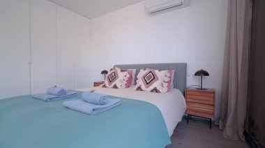 Modern yatak odasının içinde şık yastıklar, yumuşak katlanmış havlular, lambalı bir komodin ve minimalist estetik bir yatak odası var.