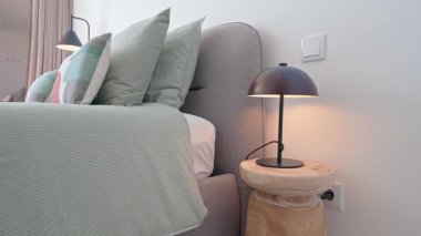Pastel yeşil ve gri renkler bu modern yatak odasında rahatlatıcı bir atmosfer yaratıyor, şık yastıklı rahat bir yatak ve doğal ahşap bir taburede minimalist bir yatak başı lambası.
