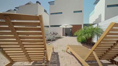 Güneş ışığı, sakin bir avludaki çakılların üzerine yerleştirilmiş iki boş ahşap iskemleyi aydınlatıyor. Konut kompleksi içindeki modern beyaz evlerin manzarasını sunuyor.