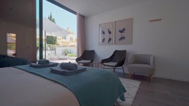 Özel bir verandaya bakan geniş pencereli rahat modern otel odası, yatağın üzerinde katlanmış havlular sergileniyor, misafirleri dinlenmeye ve rahatlamaya davet ediyor.