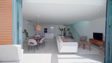 Minimalist açık planlı yaşam alanı doğal ışık banyosu, beyaz mutfak, cam merdiven ve pürüzsüz iç akım yaratan parlak mobilyalarla çağdaş tasarım sergileniyor.