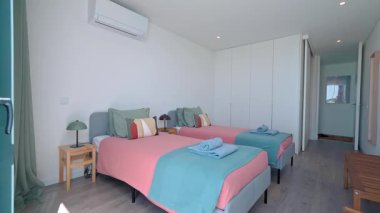 Güneş ışığı modern minimalist yatak odasını aydınlatır. Pembe ve deniz mavisi yatak örtüleriyle süslenmiş iki tek kişilik yatak. Şık geometrik desenli yastıklar ve lambalı bir komodinle tamamlanmış.