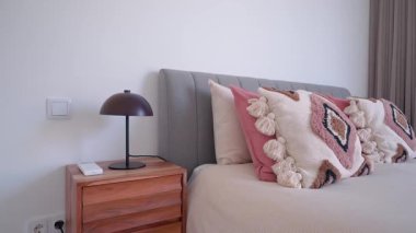 Rahat bir yatak odası, gri bir yatak başlığı, siyah lambalı ahşap bir komodin, pembe, bej ve beyaz dekoratif yastıklar, rahat ve davetkar bir atmosfer yaratıyor.
