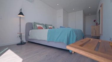 Minimalist yatak odası doğal ışıkla yıkanmış, turkuaz battaniye, gri yatak, ahşap komodin, yer lambası, gardırop ve sanat eserleri, huzur dolu çağdaş tasarım.