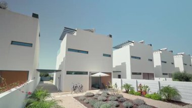 Güneş panelleri olan özdeş beyaz modern evler, açık mavi bir gökyüzü altında park halindeki bisikletlerle ve yemyeşil bahçelerle sürdürülebilir bir yerleşim alanı sergileyerek sırada duruyorlar.