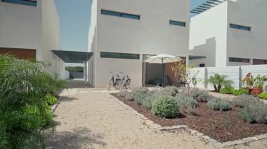 Çağdaş, çakıl yolu olan beyaz villa, park edilmiş bisikletler, veranda şemsiyesi, yemyeşil peyzaj ile çevrili huzurlu bir yerleşim ortamı yaratıyor.