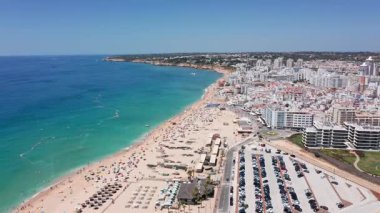 Havadan turkuaz su, park yerleri, düzenli güneşli yataklar ve şemsiyeli kumlu sahil beldesi, kıyı şeridini gezen nazik dalgalar, midrise otelleri ve gezinti güvertesi, parlak yaz gökyüzü.