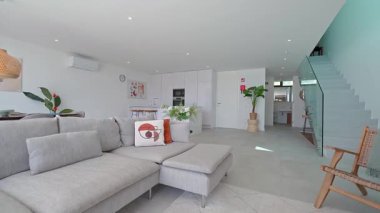 Doğal ışık, arka planda şık yastıklar ve modern bir mutfakla tamamlanmış rahat gri bir kanepenin yer aldığı geniş bir oturma odasını aydınlatarak sakin ve davetkar bir atmosfer yaratıyor.