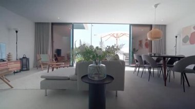 Güneş ışığı şık, çağdaş bir oturma odasını aydınlatıyor, bir yemek alanını gösteriyor, rahat bir oturma odası, ve açık cam kapılardan havuzu ve şemsiyesi olan bir teras manzarası.