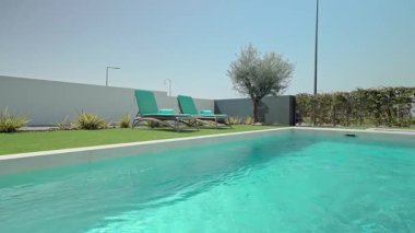 Turkuaz su, modern bir yüzme havuzunda, iki boş güneş yatağının, bir zeytin ağacının ve yeşil bir çitin yanında, huzur verici ve rahatlatıcı bir yaz atmosferi sunar.