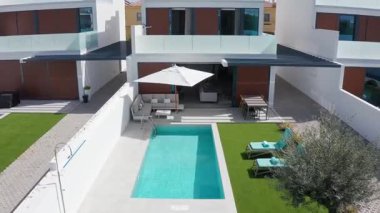 Beyaz modern tatil villası, özel yüzme havuzu, şemsiyeli veranda, masa ve sandalyeler ve sunyataklar suni çimlerin üzerinde döner manzaralı.