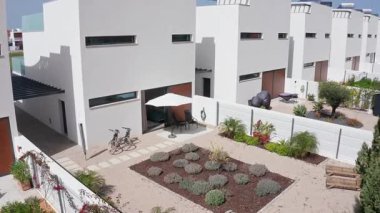 Küçük bahçeli modern beyaz evler, veranda şemsiyesi yavaş yavaş açılıyor, fayanslı bir yolda park edilmiş iki bisiklet ve bahçeleri ayıran beyaz bir çit.
