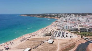 Sahil manzarası, uzak şehir manzarası yakınlarındaki kıyı şeridi, kumlu tepelerin berrak mavi gölle buluştuğu sakin su ve şemsiyeli kıyı manzarası.