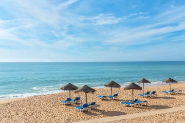 Güneşin öptüğü kumlu sahil sandalyeleri sazlı şemsiyelerin altında sergileniyor, sakin okyanus dalgaları ve berrak mavi gökyüzü tarafından rahatlatılıyor..