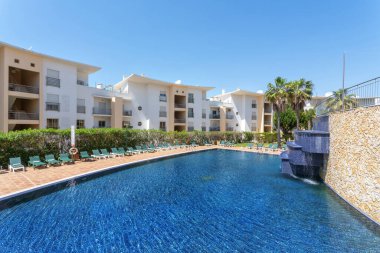 Ziyaretçiler bir tatil köyündeki havuz kenarında huzurlu bir günün tadını çıkarıyorlar. Parlak mavi su, güneşli bir öğleden sonra çevresindeki yeşillik ve modern mimariyle çelişir..