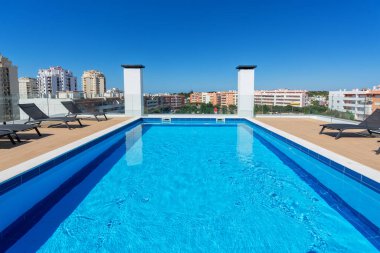 Canlı bir çatı havuzu güneşin altında parıldıyor, şezlonglarla çevrili, şehir manzarası ve parlak mavi gökyüzü..