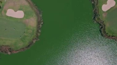 Ağaçlarla çerçevelenmiş, parıldayan gölün her iki tarafında yeşil çim yolları ve kum sığınakları olan havadan aşağı golf sahası manzarası. Spor manzarası kavramı ve doğa ile uyum