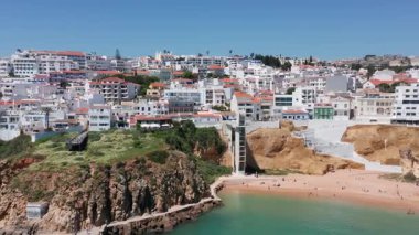Albufeira 'daki Praia dos Pescadores üzerinde sağdan sola, plaj asansörünün kumlu sahili ve yoğun tepe kenti mimarisi erişilebilir şehir plaj turizmi kavramını vurgulayan yan insansız hava aracı hareketi